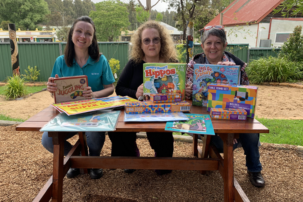 Caroline Odgers from GIVIT, sitting and smiling for a picture with Janine Hutton from Campbell Page and Julie Nichols from Mogo Aboriginal Preschool. They're holding books and games for kids